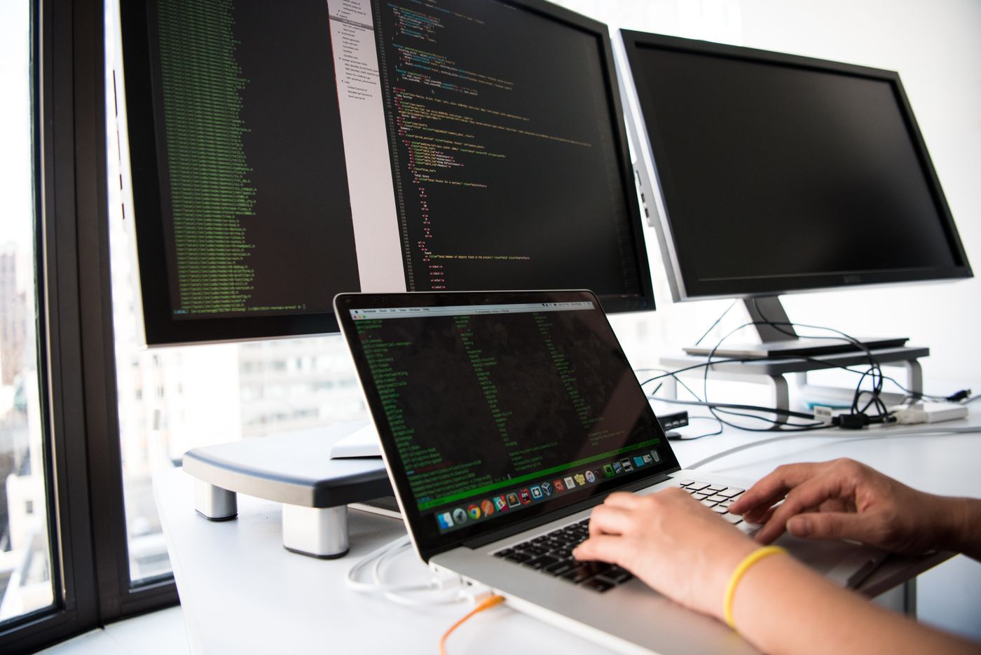 Hands typing on a laptop with code displayed on monitors